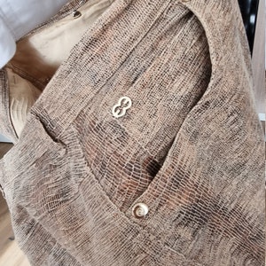 May include: A tan and brown textured fabric garment with gold-colored buttons and a decorative emblem. The garment has a pocket and a textured pattern, suggesting a reptile skin design.