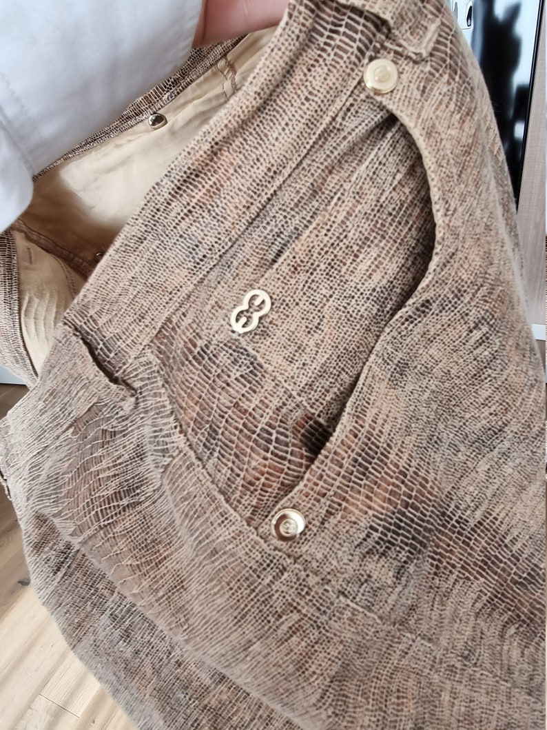 May include: A tan and brown textured fabric garment with gold-colored buttons and a decorative emblem. The garment has a pocket and a textured pattern, suggesting a reptile skin design.