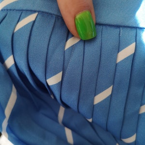 May include: Close-up of a blue and white striped fabric with pleats. The fabric has a smooth texture and is folded in a way that creates a series of vertical lines. A fingernail with bright green nail polish is visible in the upper right corner of the image.