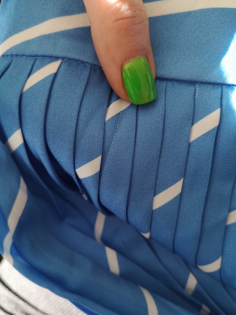 May include: Close-up of a blue and white striped fabric with pleats. The fabric has a smooth texture and is folded in a way that creates a series of vertical lines. A fingernail with bright green nail polish is visible in the upper right corner of the image.