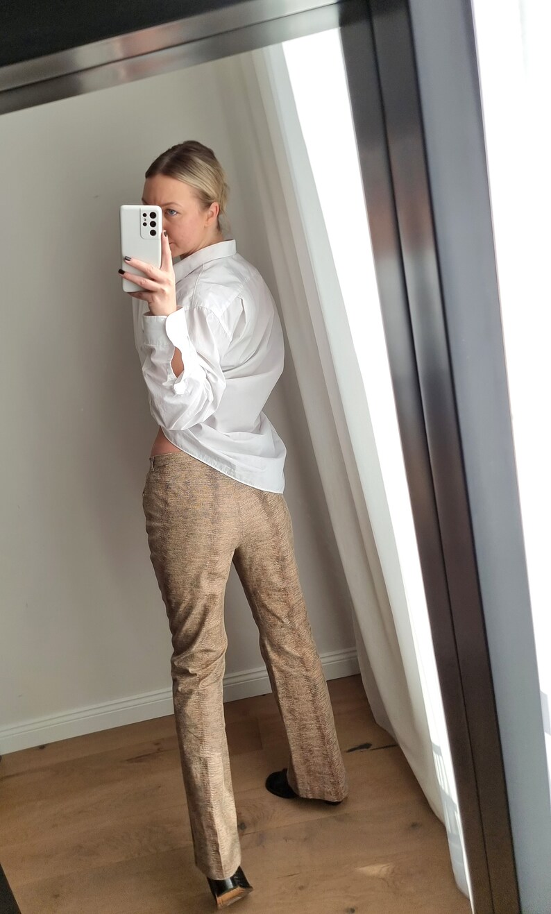 May include: A white button-down shirt and brown patterned pants are reflected in a mirror. The pants have a subtle textured design. The outfit is paired with black shoes. The image is taken indoors.