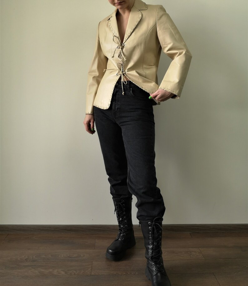 May include: A cream-colored leather jacket with a lace-up front and a pointed collar. The jacket has a fitted silhouette and is worn with black jeans and black boots.