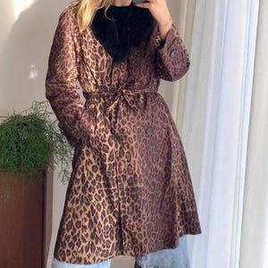 May include: A brown leopard print trench coat with a black faux fur collar. The coat has a belted waist, button closures, and long sleeves. The coat is knee-length and made of a lightweight material.