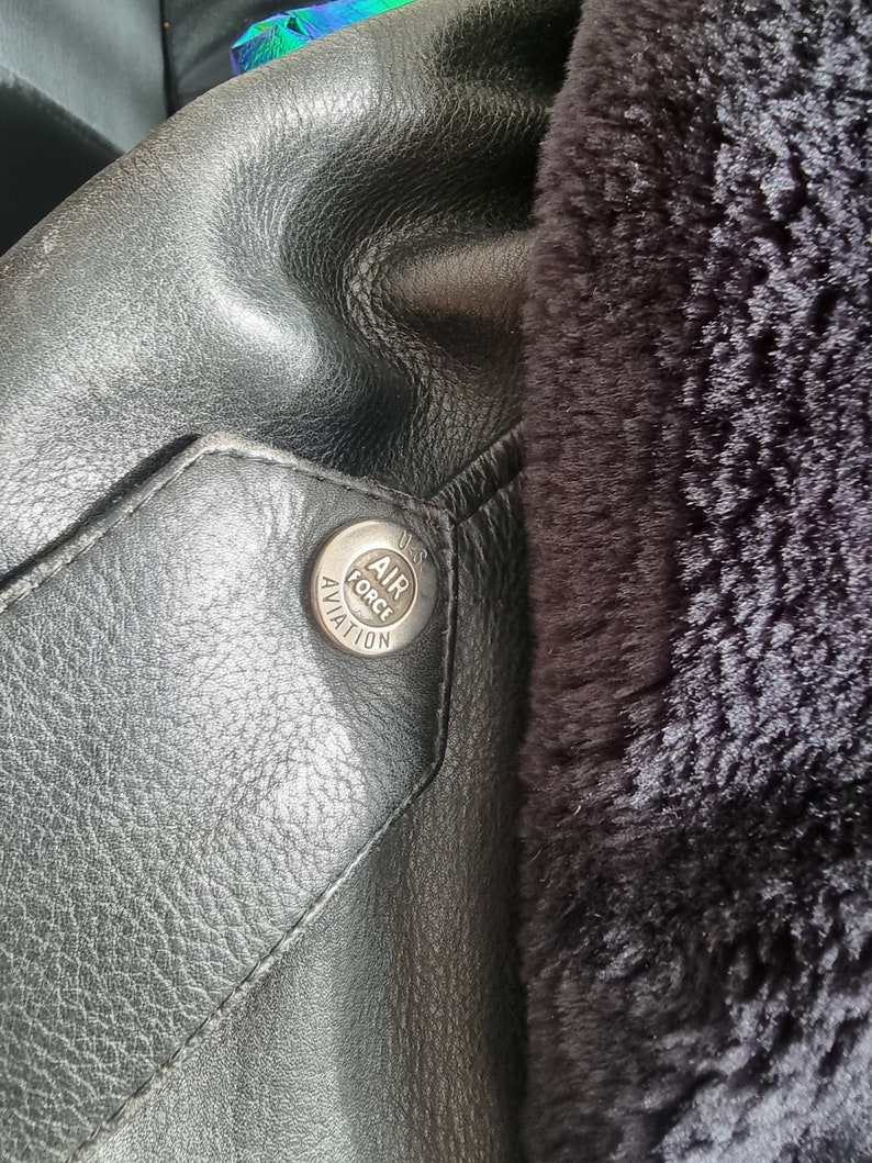 Op de afbeelding: Close-up van een zwart leren jack met een bontvoering. Een zilveren knoop met de tekst "U.S. AIR FORCE AVIATION" is zichtbaar.