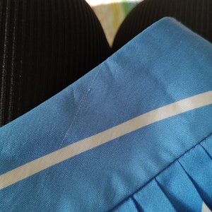 May include: Close-up of a blue and white striped skirt with a white stripe running horizontally across the fabric. The skirt has pleats and is made of a smooth, silky material.