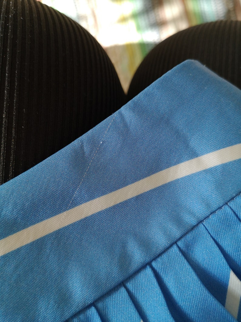 May include: Close-up of a blue and white striped skirt with a white stripe running horizontally across the fabric. The skirt has pleats and is made of a smooth, silky material.