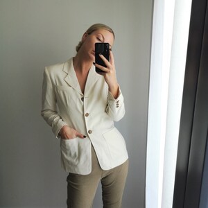 May include: A white linen blazer with a button closure and two pockets. The blazer is worn over a pair of khaki pants.
