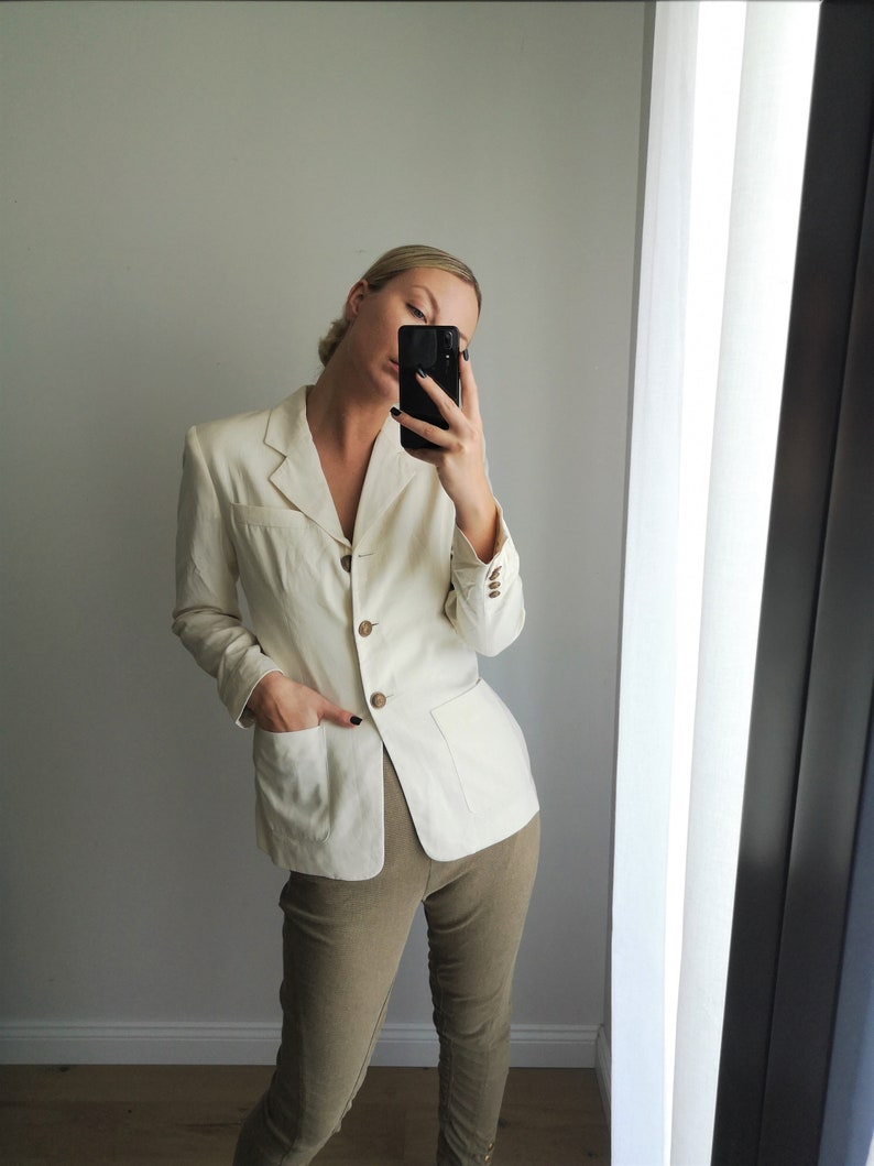 May include: A white linen blazer with a button closure and two pockets. The blazer is worn over a pair of khaki pants.