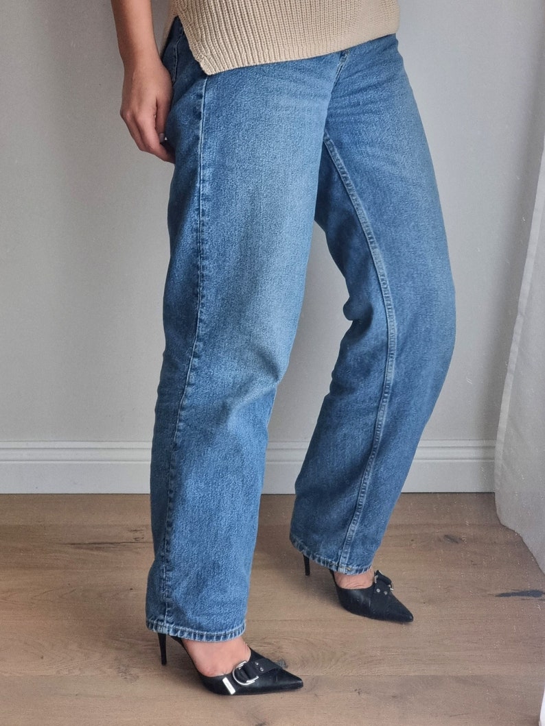 May include: A person wearing a pair of blue denim jeans with a loose fit and black heeled shoes with a buckle detail.