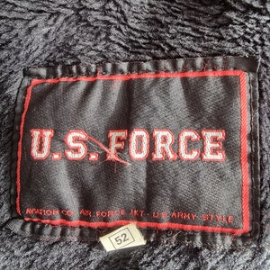 Op de afbeelding: Een zwart label met rode stiksels en de tekst "U.S. FORCE" in wit. Onder de tekst staat de tekst "AVIATION CO. AIR FORCE JKT - U.S. ARMY STYLE". Een kleiner wit label met het nummer "52" is bevestigd aan de onderkant van het grotere label.