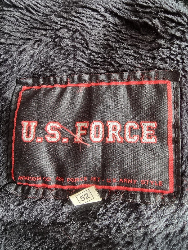 Op de afbeelding: Een zwart label met rode stiksels en de tekst "U.S. FORCE" in wit. Onder de tekst staat de tekst "AVIATION CO. AIR FORCE JKT - U.S. ARMY STYLE". Een kleiner wit label met het nummer "52" is bevestigd aan de onderkant van het grotere label.