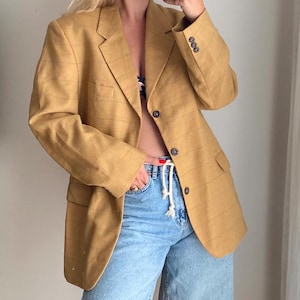 May include: Mustard yellow blazer with a plaid pattern. The blazer has a notched lapel, two front pockets, and three buttons on each cuff. The model is wearing light blue jeans.