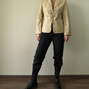 May include: A cream-colored leather jacket with a lace-up front and a pointed collar. The jacket is worn by a person with black jeans and black boots.