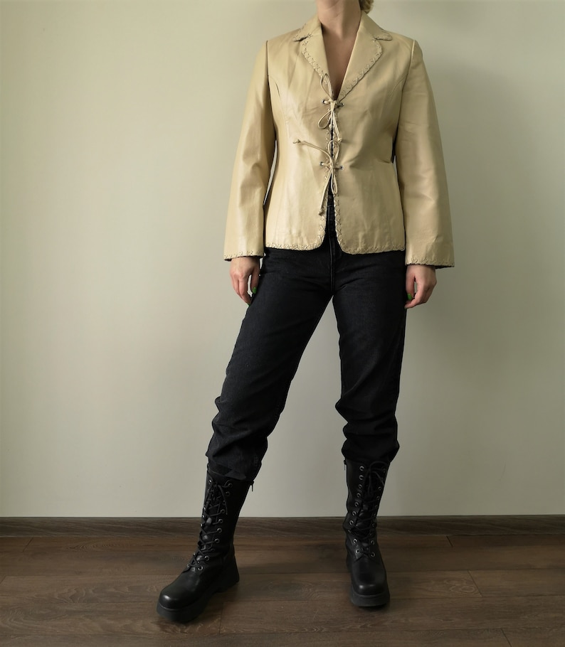 May include: A cream-colored leather jacket with a lace-up front and a pointed collar. The jacket is worn by a person with black jeans and black boots.