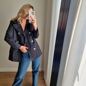 May include: A navy blue double-breasted peacoat with large silver buttons. The coat is worn over blue jeans.
