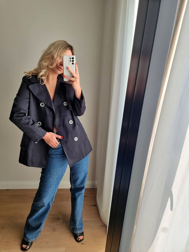 May include: A navy blue double-breasted peacoat with large silver buttons. The coat is worn over blue jeans.
