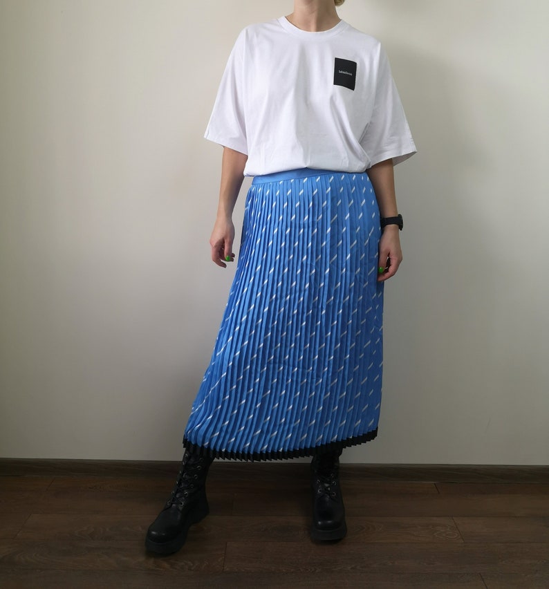 May include: A blue pleated skirt with a white geometric pattern. The skirt has a black waistband and a black trim at the hem.