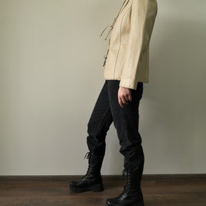 May include: A cream-colored leather jacket with a lace-up front and a fitted silhouette. The jacket is worn with black jeans and black lace-up boots.