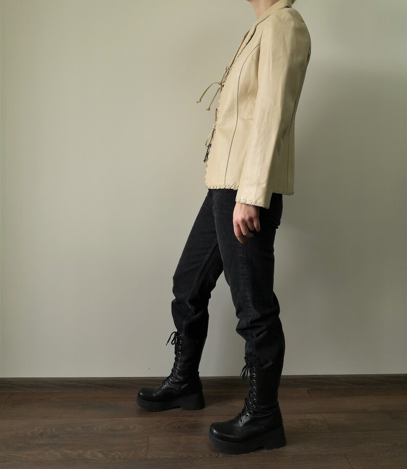 May include: A cream-colored leather jacket with a lace-up front and a fitted silhouette. The jacket is worn with black jeans and black lace-up boots.