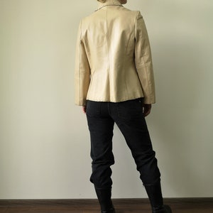 May include: A beige leather blazer with decorative stitching. The blazer has a notched collar and long sleeves. The model is wearing black jeans and black boots.