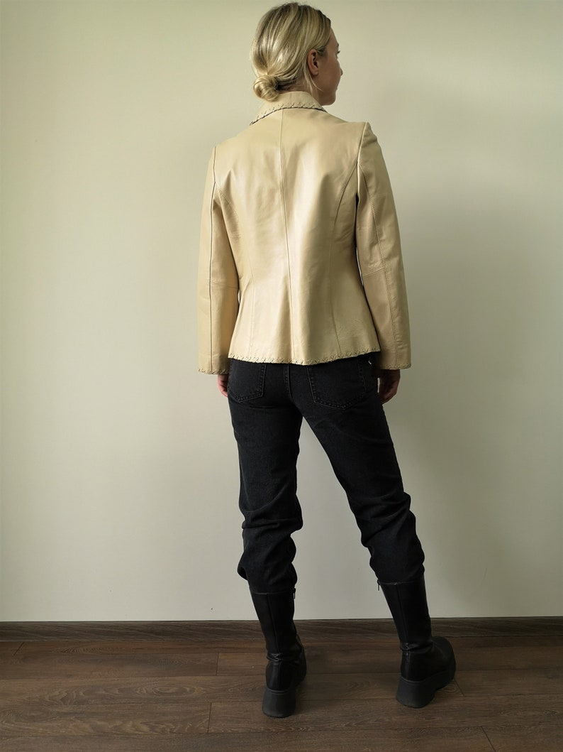May include: A beige leather blazer with decorative stitching. The blazer has a notched collar and long sleeves. The model is wearing black jeans and black boots.