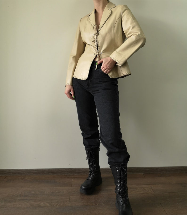 May include: A cream-colored leather jacket with a lace-up front and a fitted silhouette. The jacket has a notched collar and long sleeves. The model is wearing black jeans and black boots.