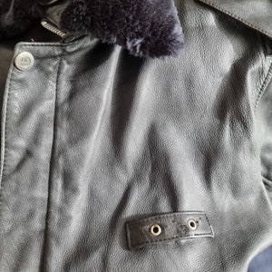 Op de afbeelding: Zwart leren jack met een bontkraag en een ritssluiting. Het jack heeft een klein patch met de tekst "U.S. AIR FORCE" aan de linkerkant van de borst.