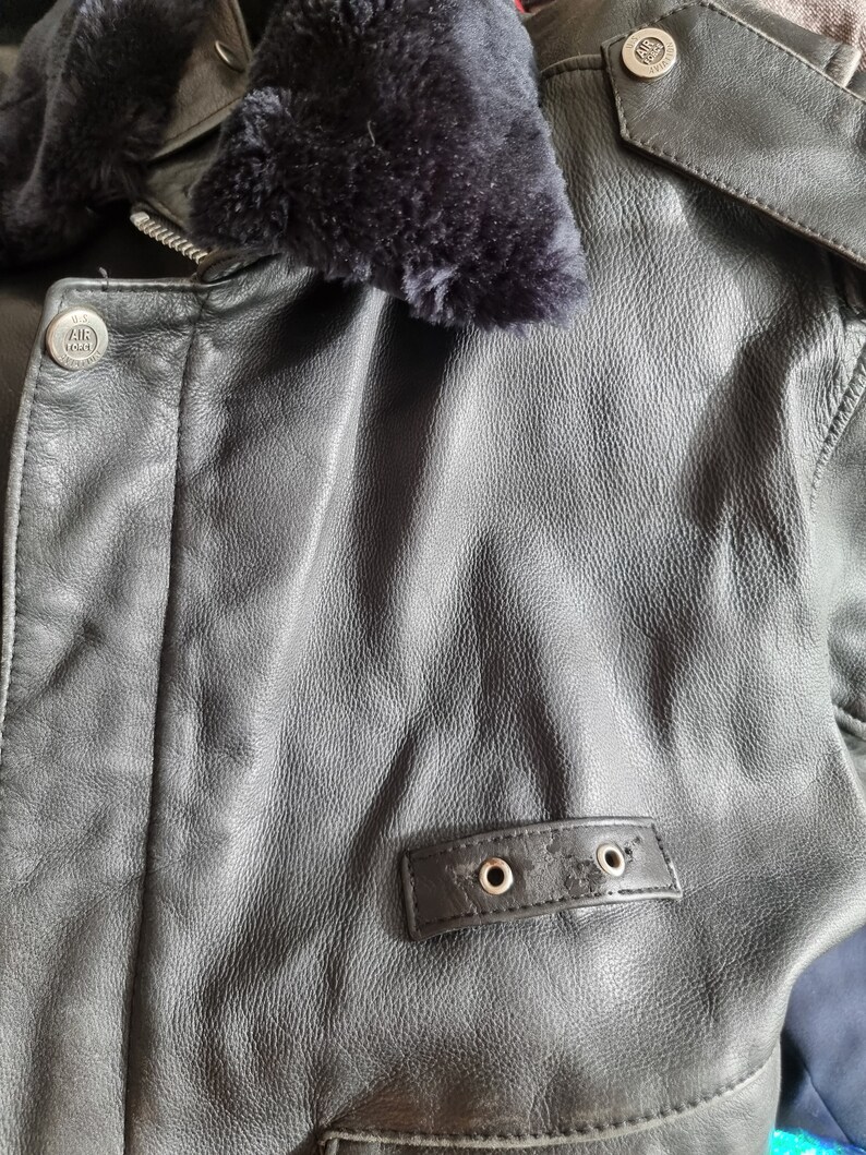 Op de afbeelding: Zwart leren jack met een bontkraag en een ritssluiting. Het jack heeft een klein patch met de tekst "U.S. AIR FORCE" aan de linkerkant van de borst.