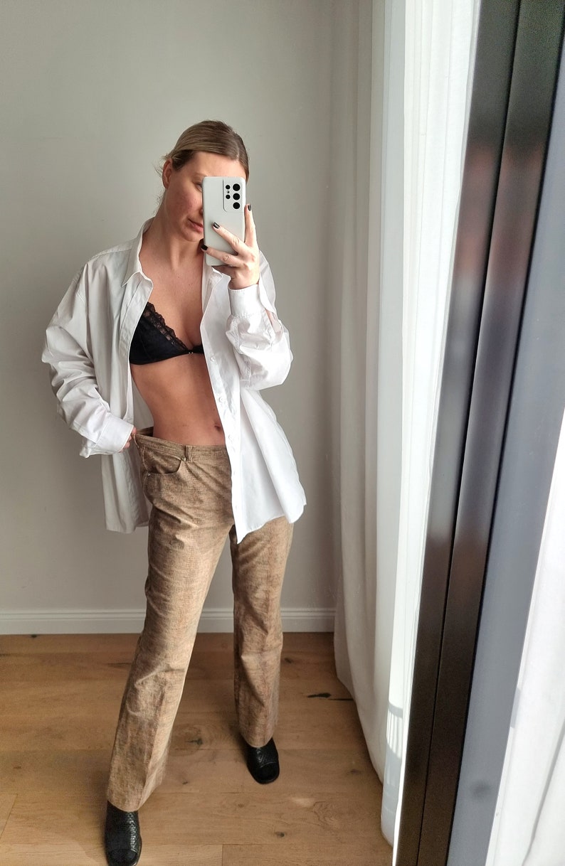 May include: A white button-down shirt, black lace bra, and tan pants are worn. The pants have a textured appearance. Black shoes complete the outfit. The person is holding a phone up to take a picture in front of a mirror.