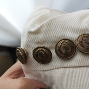 May include: Close-up of a white fabric with four gold-toned buttons. Each button has a raised design with a laurel wreath and the letters "G.P."