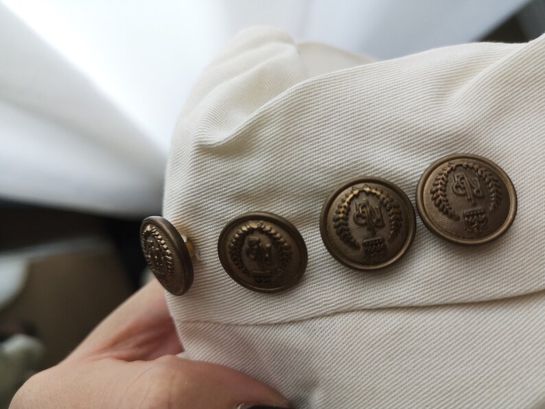 May include: Close-up of a white fabric with four gold-toned buttons. Each button has a raised design with a laurel wreath and the letters "G.P."