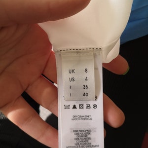 May include: White clothing label with black text and symbols. The label shows size information in UK, US, F, and I sizes. The label also includes care instructions, fabric content, and the brand name "Victoria Beckham".