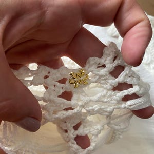 May include: Close-up of a white crocheted fabric with a gold metal logo featuring the letters "W" and "M" intertwined.