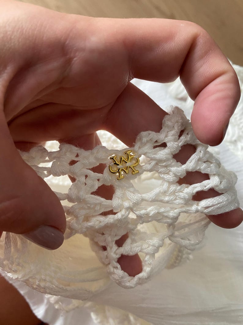 May include: Close-up of a white crocheted fabric with a gold metal logo featuring the letters "W" and "M" intertwined.