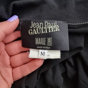 May include: Black Jean Paul Gaultier label with white stitching. The label reads "Jean Paul Gaultier" and "MailIe" with "Made in Italy" below. A white size tag with a black "M" is attached to the label.
