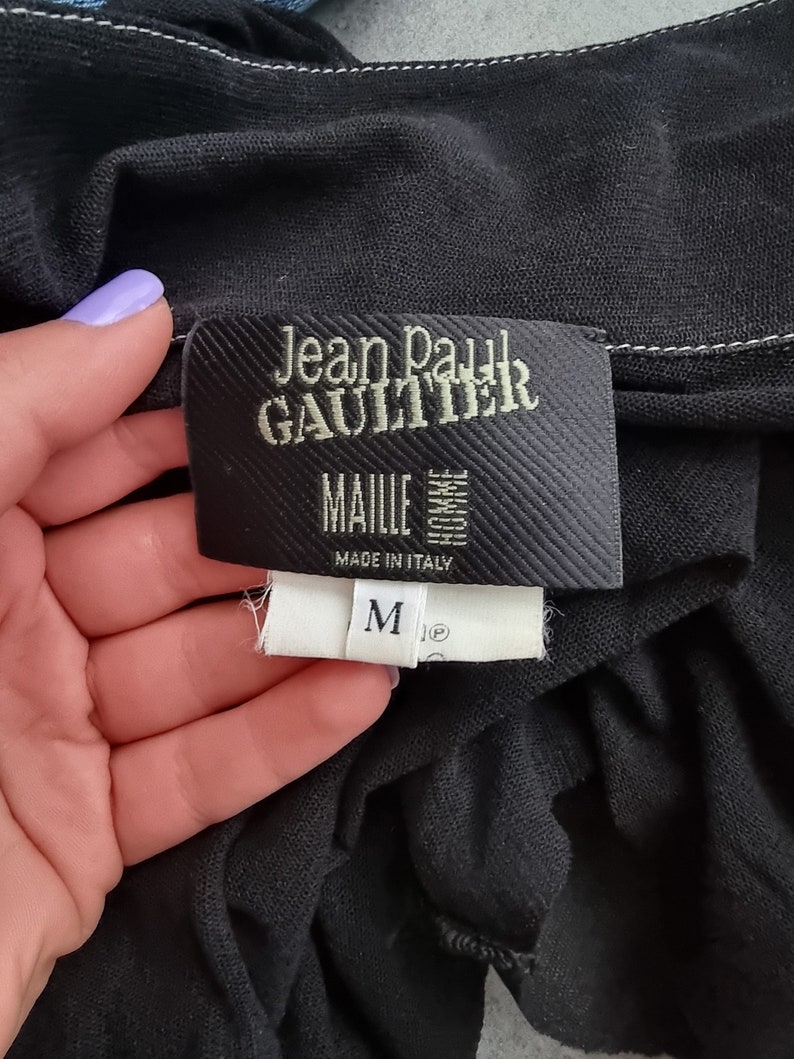 May include: Black Jean Paul Gaultier label with white stitching. The label reads "Jean Paul Gaultier" and "MailIe" with "Made in Italy" below. A white size tag with a black "M" is attached to the label.