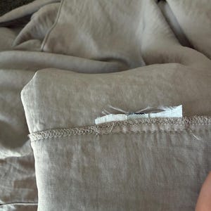 Puede incluir: Primer plano de una tela de color beige claro, posiblemente lino o algod&oacute;n, con un borde doblado y costuras visibles. Una peque&ntilde;a etiqueta blanca est&aacute; adherida a la tela. La tela tiene un aspecto suave y ligeramente arrugado.