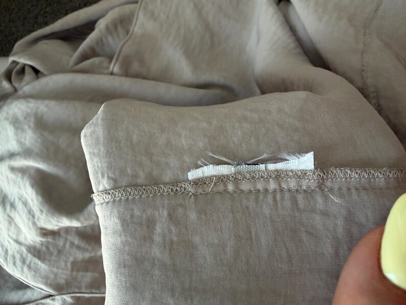 Puede incluir: Primer plano de una tela de color beige claro, posiblemente lino o algod&oacute;n, con un borde doblado y costuras visibles. Una peque&ntilde;a etiqueta blanca est&aacute; adherida a la tela. La tela tiene un aspecto suave y ligeramente arrugado.