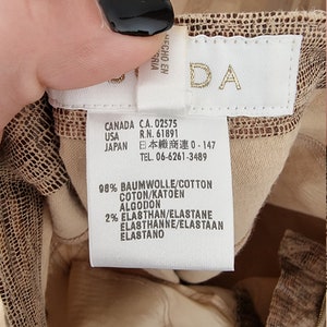 May include: A close-up of a clothing tag with the brand name "DA" in gold lettering. The tag includes information about the garment's composition: 98% cotton and 2% elastane. The tag also lists the country of origin as Canada, USA, and Japan.
