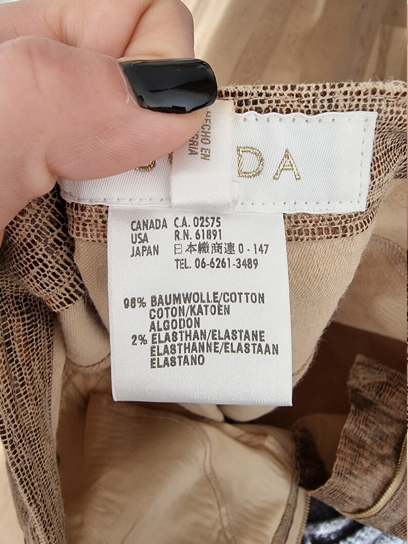 May include: A close-up of a clothing tag with the brand name "DA" in gold lettering. The tag includes information about the garment's composition: 98% cotton and 2% elastane. The tag also lists the country of origin as Canada, USA, and Japan.