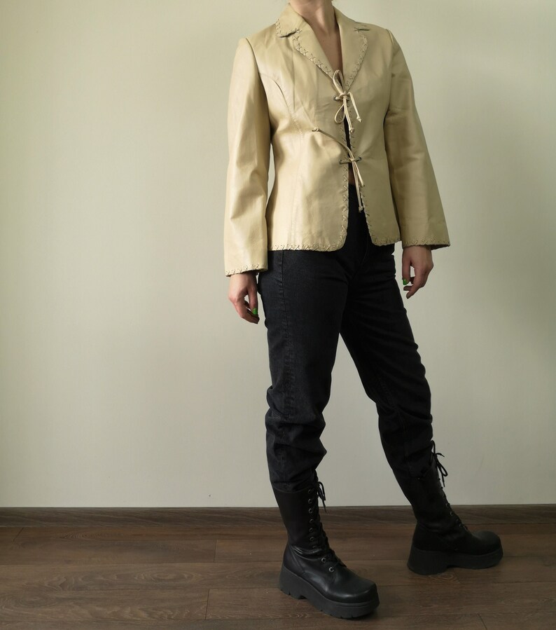 May include: A cream-colored leather jacket with a lace-up front and a fitted silhouette. The jacket has a collar and long sleeves.