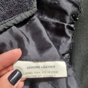 Op de afbeelding: Een zwart leren jack met een wit label met de tekst "GENUINE LEATHER, LINING 100% POLYESTER, SPECIALIST CLEANER."