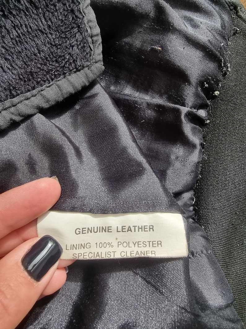 Op de afbeelding: Een zwart leren jack met een wit label met de tekst "GENUINE LEATHER, LINING 100% POLYESTER, SPECIALIST CLEANER."