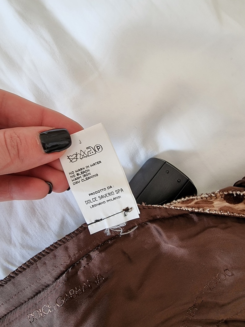 Puede incluir: Una etiqueta de cuidado blanca con texto negro y s&iacute;mbolos. La etiqueta dice "NO WASH IN WATER, NO BLEACH, WARM IRON, DRY CLEANING". Tambi&eacute;n dice "PRODOTTO DA: DOLCE SAVERIO SPA LEGNANO IMILANO".