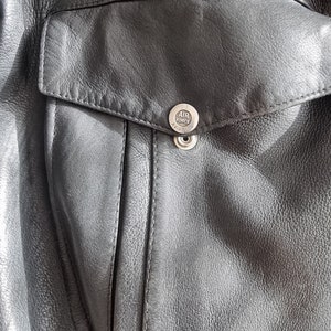 Op de afbeelding: Close-up van een zwart leren jack met een zak en een zilveren knoop met de tekst "U.S. AIR FORCE AVIATION".