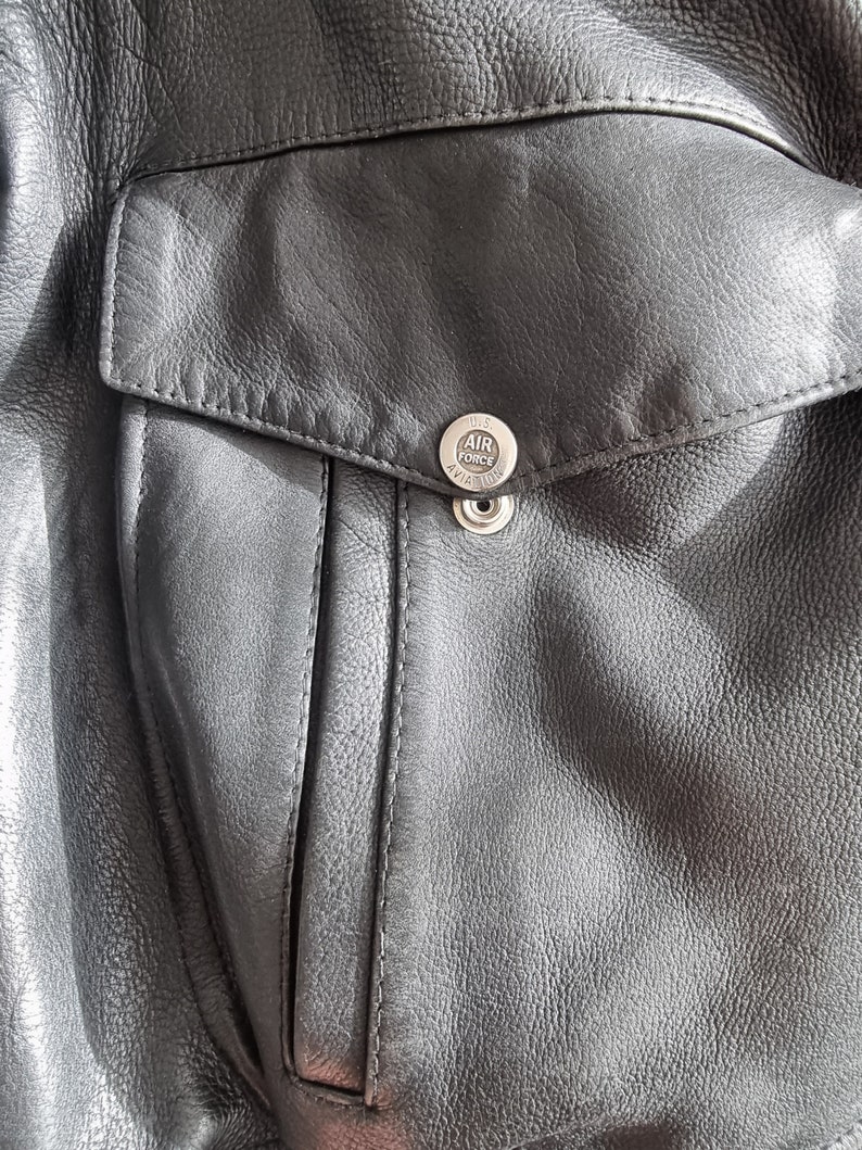 Op de afbeelding: Close-up van een zwart leren jack met een zak en een zilveren knoop met de tekst "U.S. AIR FORCE AVIATION".