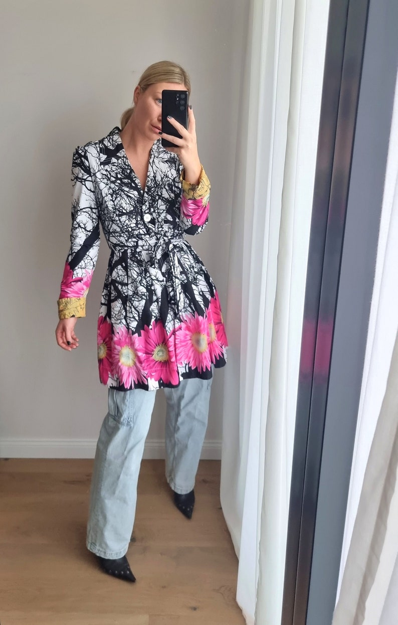 May include: A white and black floral print jacket with a pink and yellow floral trim. The jacket has a cinched waist and a long, flowing skirt. The model is wearing light blue jeans and black heeled shoes.