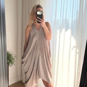 Puede incluir: Un vestido halter de color topo claro con un dise&ntilde;o drapeado. El vestido tiene un escote en V y un dobladillo asim&eacute;trico. La persona de la imagen lleva sandalias negras. El vestido est&aacute; hecho de una tela suave y fluida.
