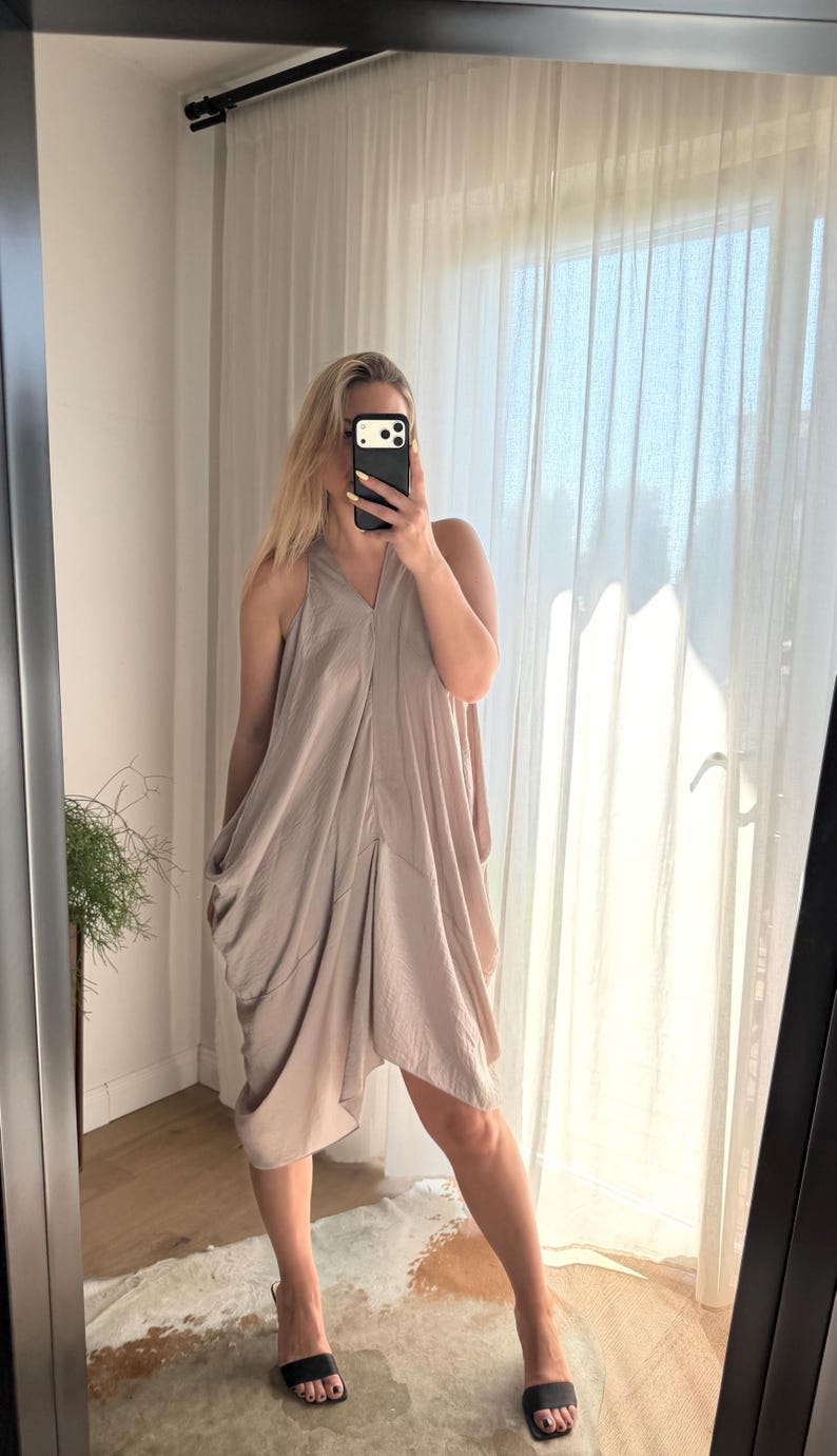 Puede incluir: Un vestido halter de color topo claro con un dise&ntilde;o drapeado. El vestido tiene un escote en V y un dobladillo asim&eacute;trico. La persona de la imagen lleva sandalias negras. El vestido est&aacute; hecho de una tela suave y fluida.