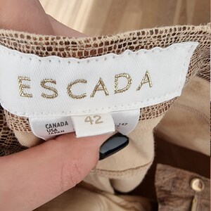 May include: Close-up of a clothing label with the brand name "ESCADA" in gold lettering. The label is white with the size "42" printed on it. The label also says "CANADA USA JAPAN". The fabric is a brown and tan snakeskin pattern.